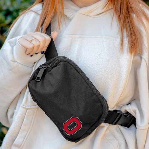A person wearing a white hoodie and a black Ohio State Buckeyes belt bag with an adjustable strap and the official red and gray block logo on the front.