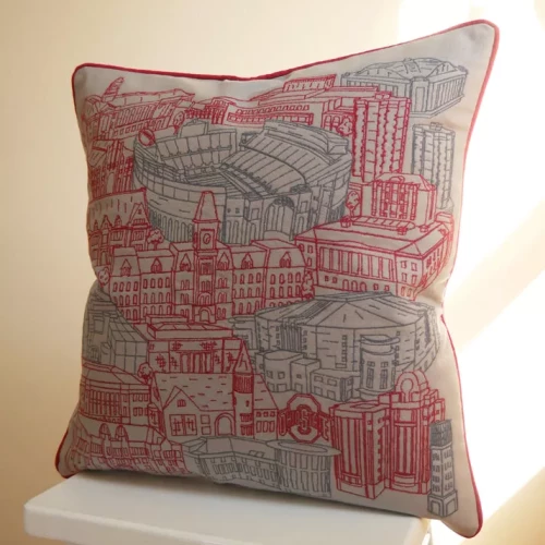An Ohio State throw pillow featuring a detailed illustration of places around campus , resting on a white wooden stool.