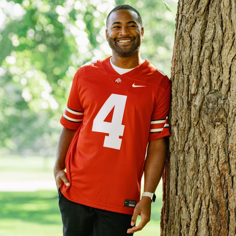 A model leans against a tree on the Oval wearing a red Ohio State #4 jersey. 