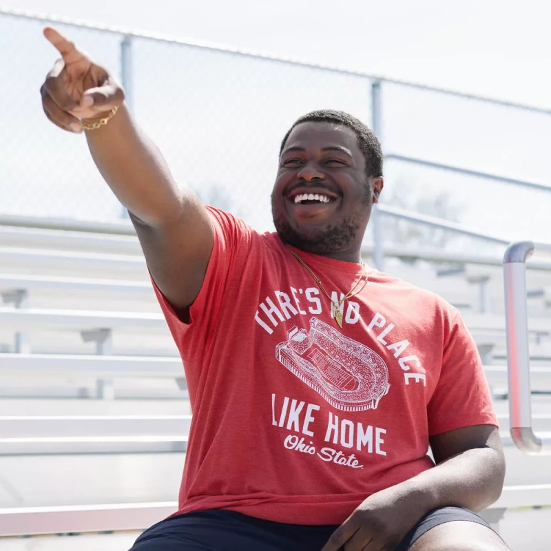 a model points to his right, as if greeting a friend. he wears a red shirt that says 